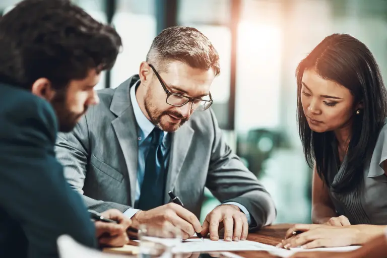 Three people in a business meeting, sitting around a table and reviewing documents. The man in the center wears glasses and a suit, and is writing.