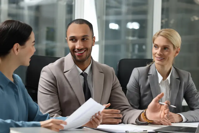 Three people in business attire have a meeting. The man in the middle is speaking, holding a pen. The woman on the left holds papers, while the woman on the right listens attentively.