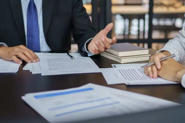 Two people are sitting at a table with documents, discussing or signing papers. One is gesturing, and there are books in the background.