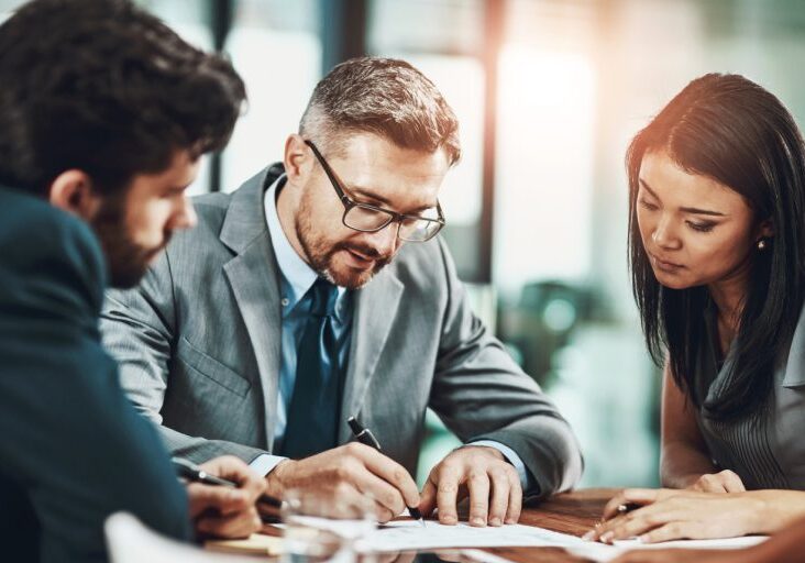 Three people in a business meeting, sitting around a table and reviewing documents. The man in the center wears glasses and a suit, and is writing.