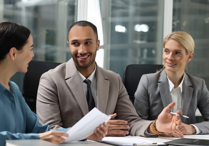 Three people in business attire have a meeting. The man in the middle is speaking, holding a pen. The woman on the left holds papers, while the woman on the right listens attentively.