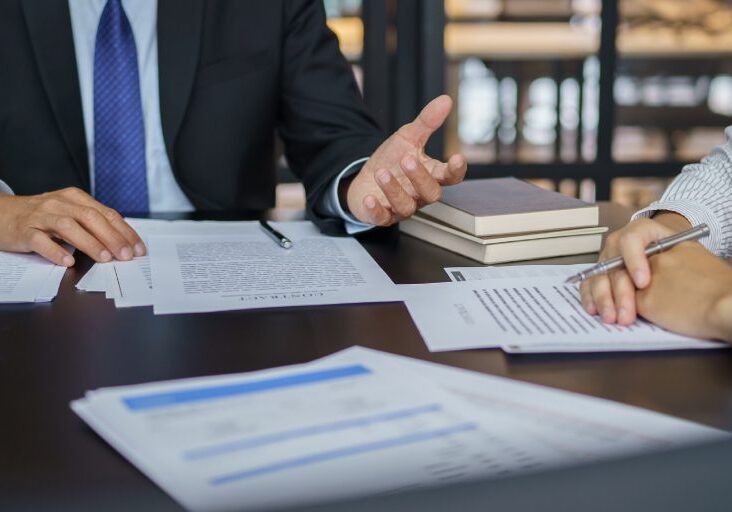 Two people are sitting at a table with documents, discussing or signing papers. One is gesturing, and there are books in the background.
